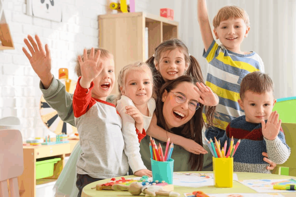 Little Antlers Childcare teacher smiling at a desk surrounded by five preschool children waving hello in a bright classroom.